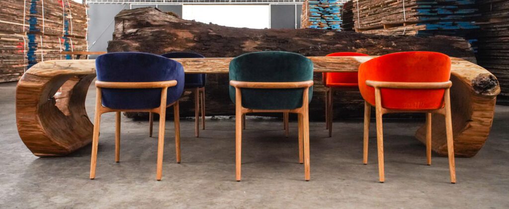 A live-edge oak slab dining table carved into a continuous loop form, paired with six velvet-upholstered chairs in blue, green, and orange, set in an industrial showroom.